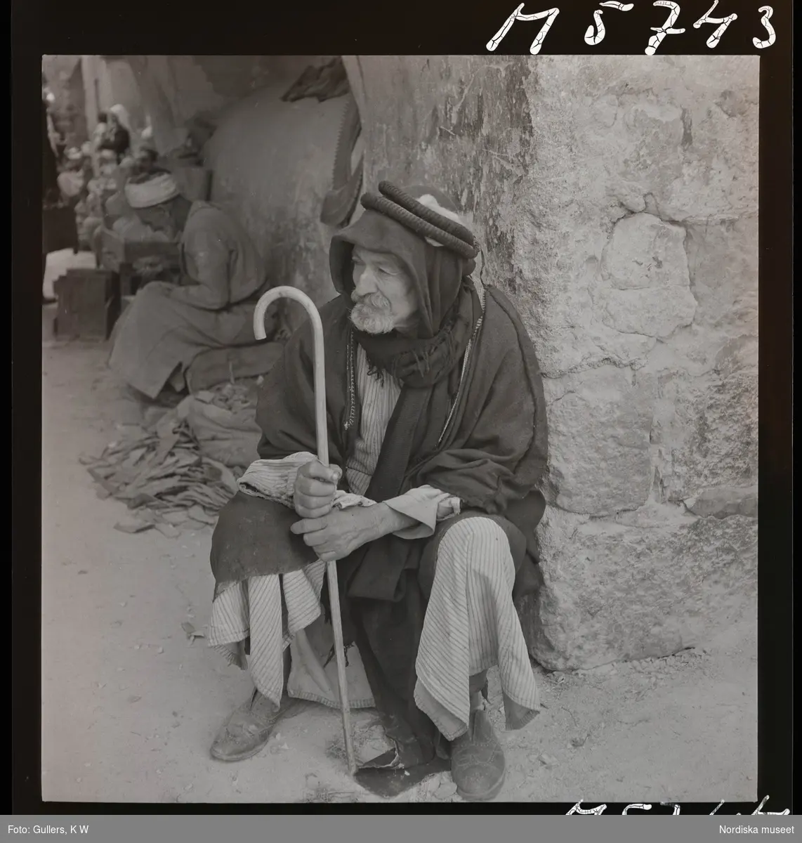 Äldre man med käpp sitter i ett gathörn.