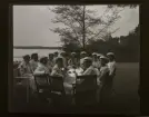 En stor grupp kvinnliga studenter klädda i vitt sitter utomhus vid ett stort bord och dricker kaffe.