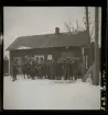 Ett jaktlag med vapen och redskap i händerna framför J. S. Anderssons skrädderi, speceri- & diversehandel.