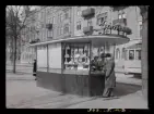 Kiosk. Strandvägen, Stockholm. En man i hatt handlar i luckan.