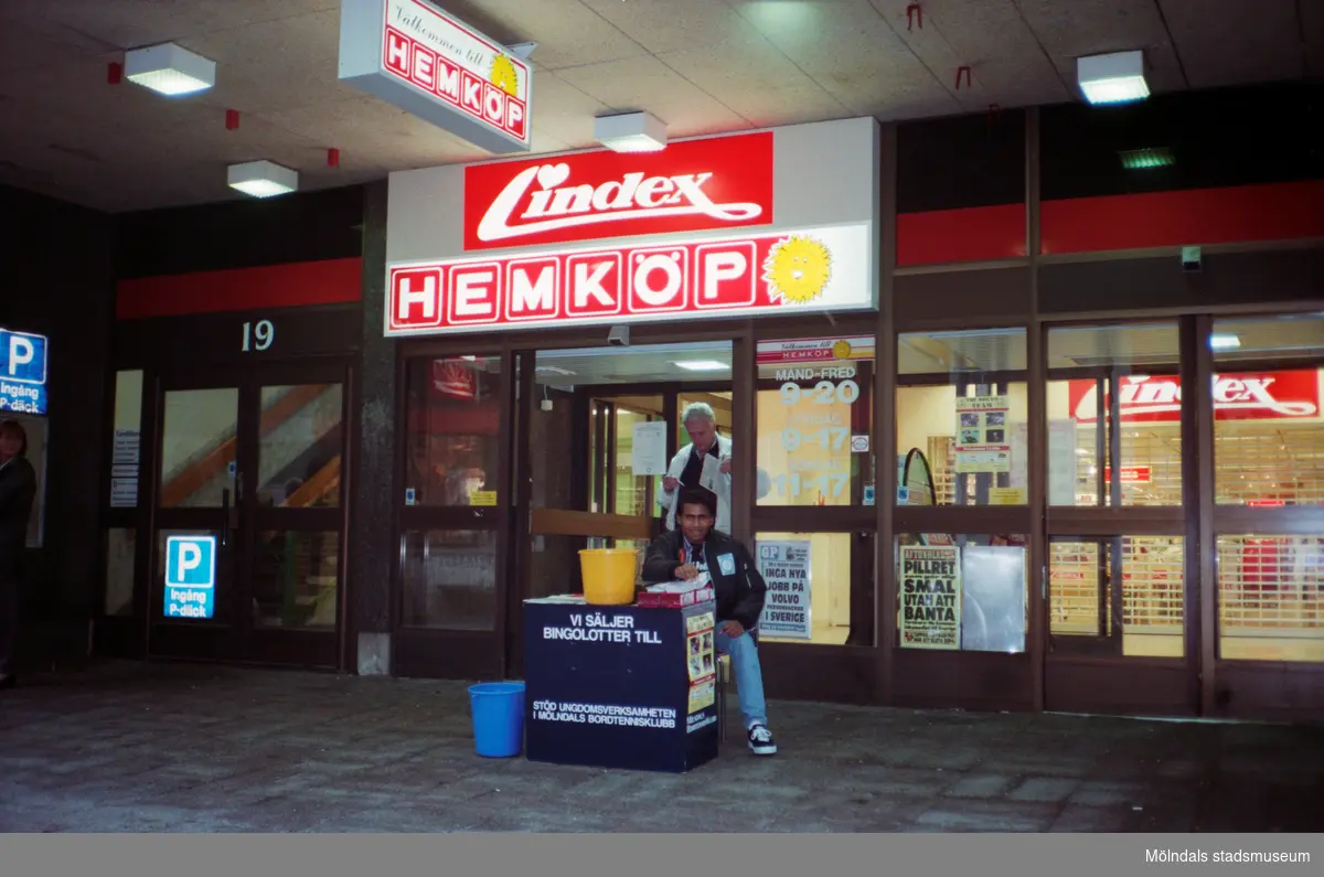 Bingolottoförsäljning på Brogatan 19 utanför Lindex och Hemköp. Mölndalsbro i dag - ett skolpedagogiskt dokumentationsprojekt på Mölndals museum under oktober 1996. 1996_1212-1226 är gjorda av högstadieelever från Kvarnbyskolan 9D, grupp 5. Se även 1996_0913-0940.