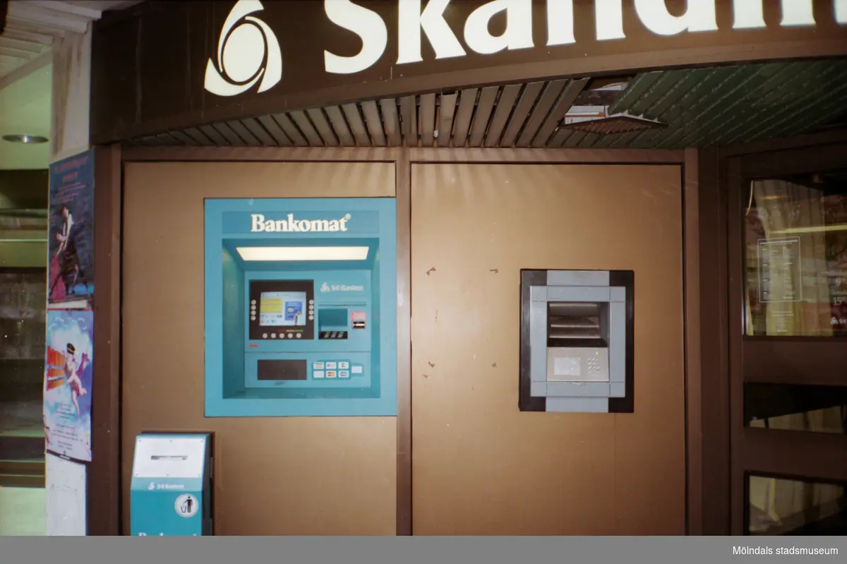 Bankomat utanför Skandinaviska Enskilda Banken på Brogatan/Nygatan. Mölndalsbro i dag - ett skolpedagogiskt dokumentationsprojekt på Mölndals museum under oktober 1996. 1996_1212-1226 är gjorda av högstadieelever från Kvarnbyskolan 9D, grupp 5. Se även 1996_0913-0940.