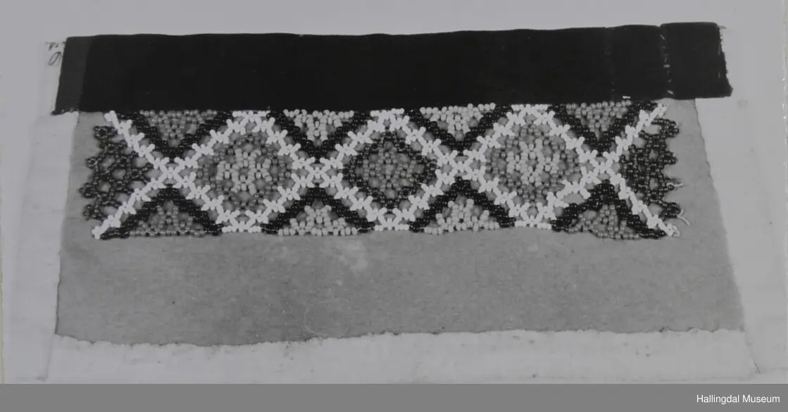 Kvit bomullslerret på baksida og bretta noko over på framsida. På frramsida raud vadmål med påsydd geometrisk mønster av små, runde glasperler. Øvst på brystduken ein svart fløyelskant.