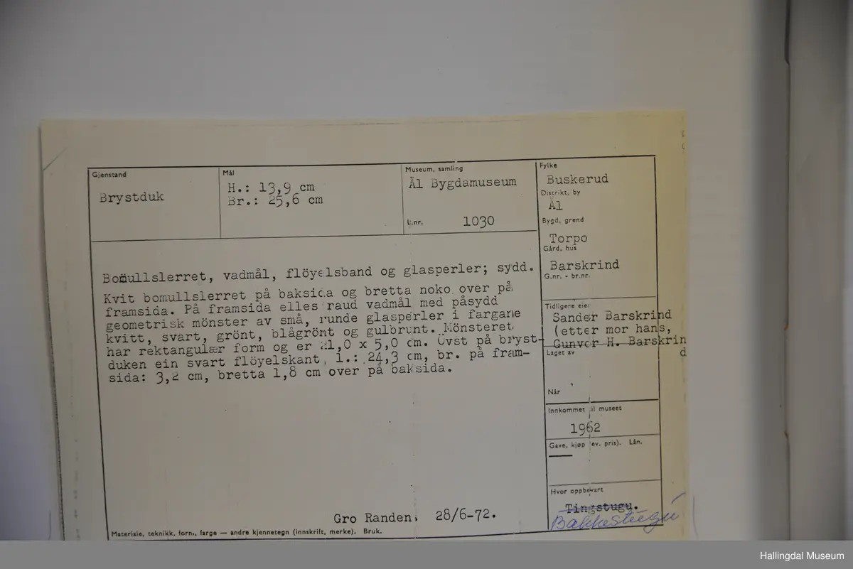 Kvit bomullslerret på baksida og bretta noko over på framsida. På frramsida raud vadmål med påsydd geometrisk mønster av små, runde glasperler. Øvst på brystduken ein svart fløyelskant.