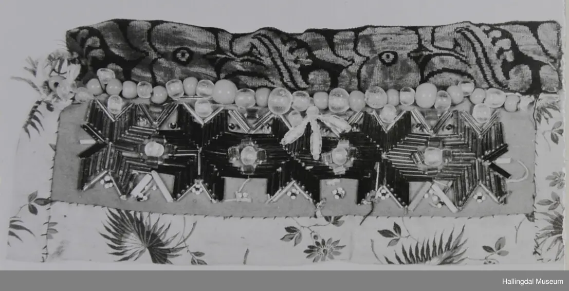 5 åttebladroser av lange glasperler, små perler rundt, over desse ei rad med store, kuleforma, blanke og lys blå perler.