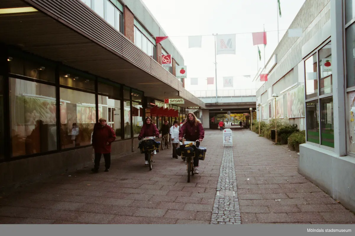 Människor som cyklar och går på Nygatan. I fonden ses bron. Mölndalsbro i dag - ett skolpedagogiskt dokumentationsprojekt på Mölndals museum under oktober 1996. 1996_1264-1282 är gjorda av högstadieelever från Åbyskolan 8A, grupp 2. Se även 1996_0913-0940, gruppbilder på klasserna 1996_1382-1405 och bilder från den färdiga utställningen 1996_1358-1381.