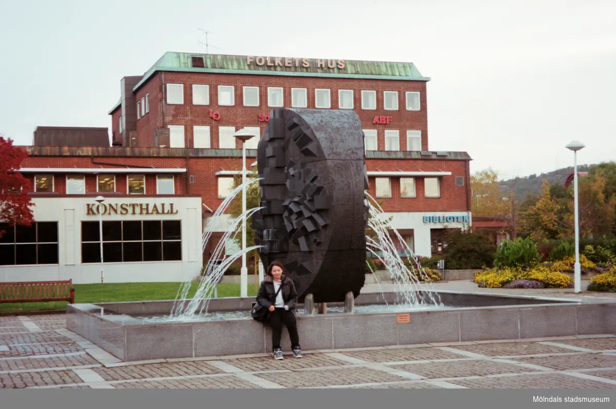 En kvinna sitter vid en fontän framför Folkets hus på Stadshusplatsen. Mölndalsbro i dag - ett skolpedagogiskt dokumentationsprojekt på Mölndals museum under oktober 1996. 1996_1320-1337 är gjorda av högstadieelever från Åbyskolan 8A, grupp 5. Se även 1996_0913-0940, gruppbilder på klasserna 1996_1382-1405 och bilder från den färdiga utställningen 1996_1358-1381.