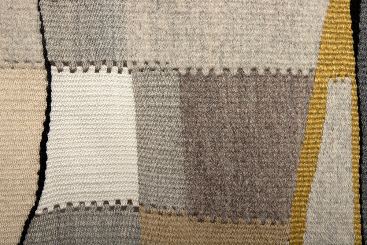 I grå, beige, gule og sorte toner vises et motiv av firkantede og trekantede fargefelt. Mellom de skrå linjene fremhever vevteknikken skillene mellom fargefeltene.