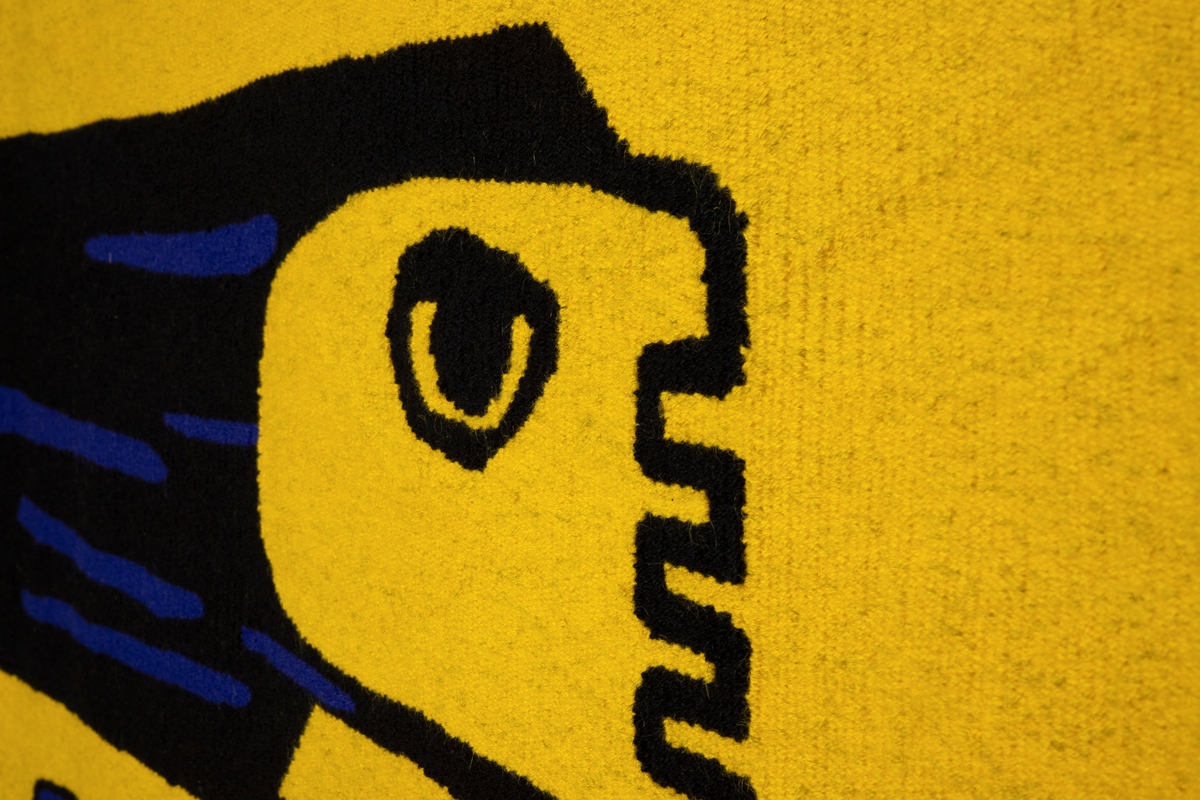 Rektangulær billedvev i sort og blålilla på gul bakgrunn. Motivet viser en figur i profil. Hodet er stillisert med enkle detaljer, og håret vises som et sort felt med blålilla detaljer stående rett ut mot venstre kant. Kroppen er bygget opp av vertikale streker som bukter seg ned mot føttene. Føttene peker rett ned. Hånda vises som heldekkende felt i sort og blålilla. Kunstnerens signatur er vevet inn som en S i det nedre venstre hjørnet. Bokstaven er som kroppen bygget opp av buktende enkeltlinjer.