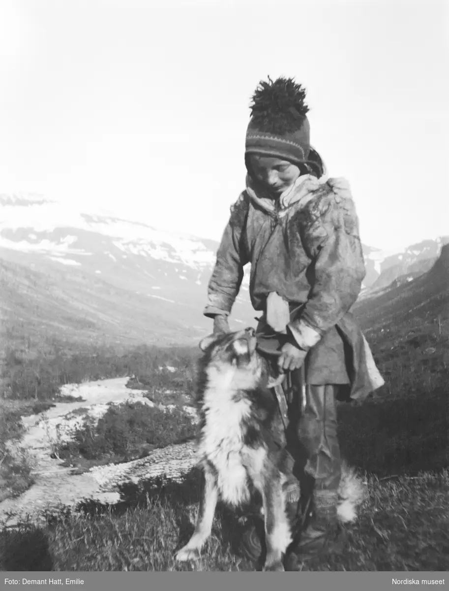 Ett barn leker med en hund (Curri), enligt bildtexten är pojken Andaras Ommas yngste son. I bakgrunden fjäll och en dalgång. Bilden ingår i en serie fotografier tagna av Emilie Demant Hatt i Sapmi mellan åren 1907 och 1916.