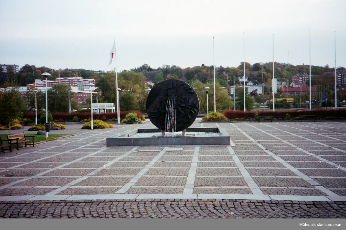 Vy österut på Stadshusplatsens fontän/skulptur och flaggstänger. Mölndalsbro i dag - ett skolpedagogiskt dokumentationsprojekt på Mölndals museum under oktober 1996. 1996_1338-1356 är gjorda av högstadieelever från Åbyskolan 8A, grupp 6. Se även 1996_0913-0940, gruppbilder på klasserna 1996_1382-1405 och bilder från den färdiga utställningen 1996_1358-1381.