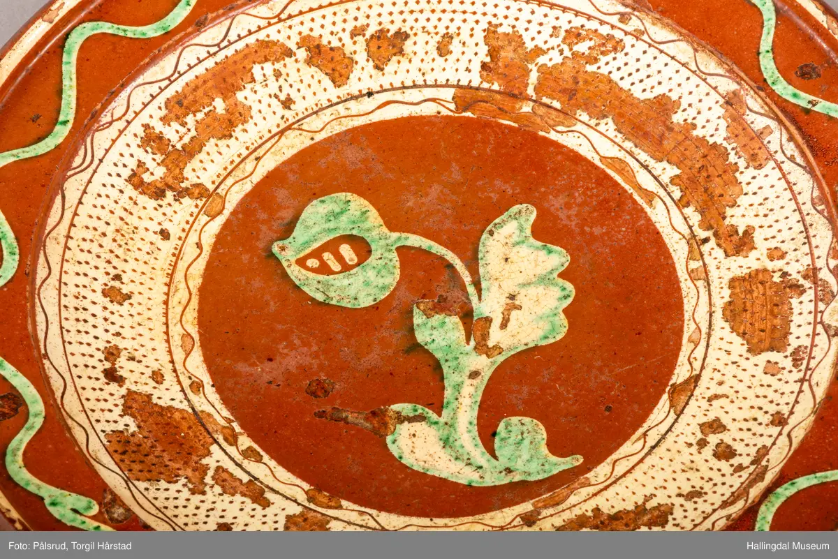 Brunt keramikkfat med dekor i grønt og hvitt. Glassert. Grønn tulipan på midten med tykk hvit bord rundt. Bølget grønn bord langs kanten med hvit tynn bord ytterst.