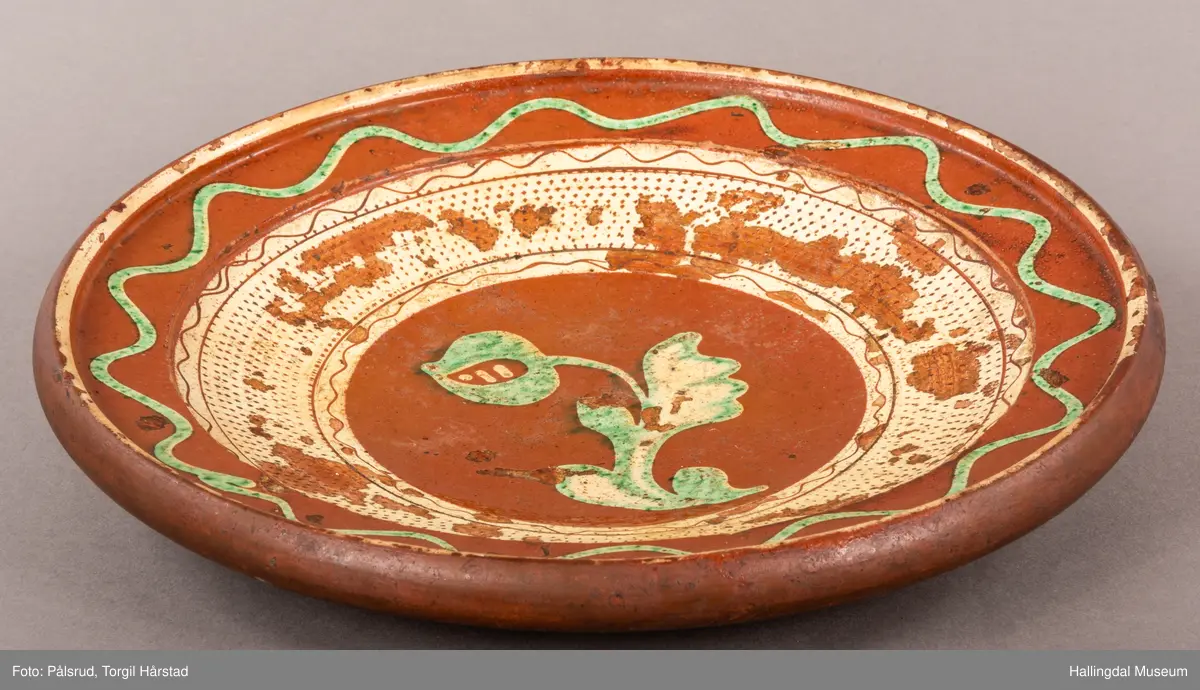 Brunt keramikkfat med dekor i grønt og hvitt. Glassert. Grønn tulipan på midten med tykk hvit bord rundt. Bølget grønn bord langs kanten med hvit tynn bord ytterst.