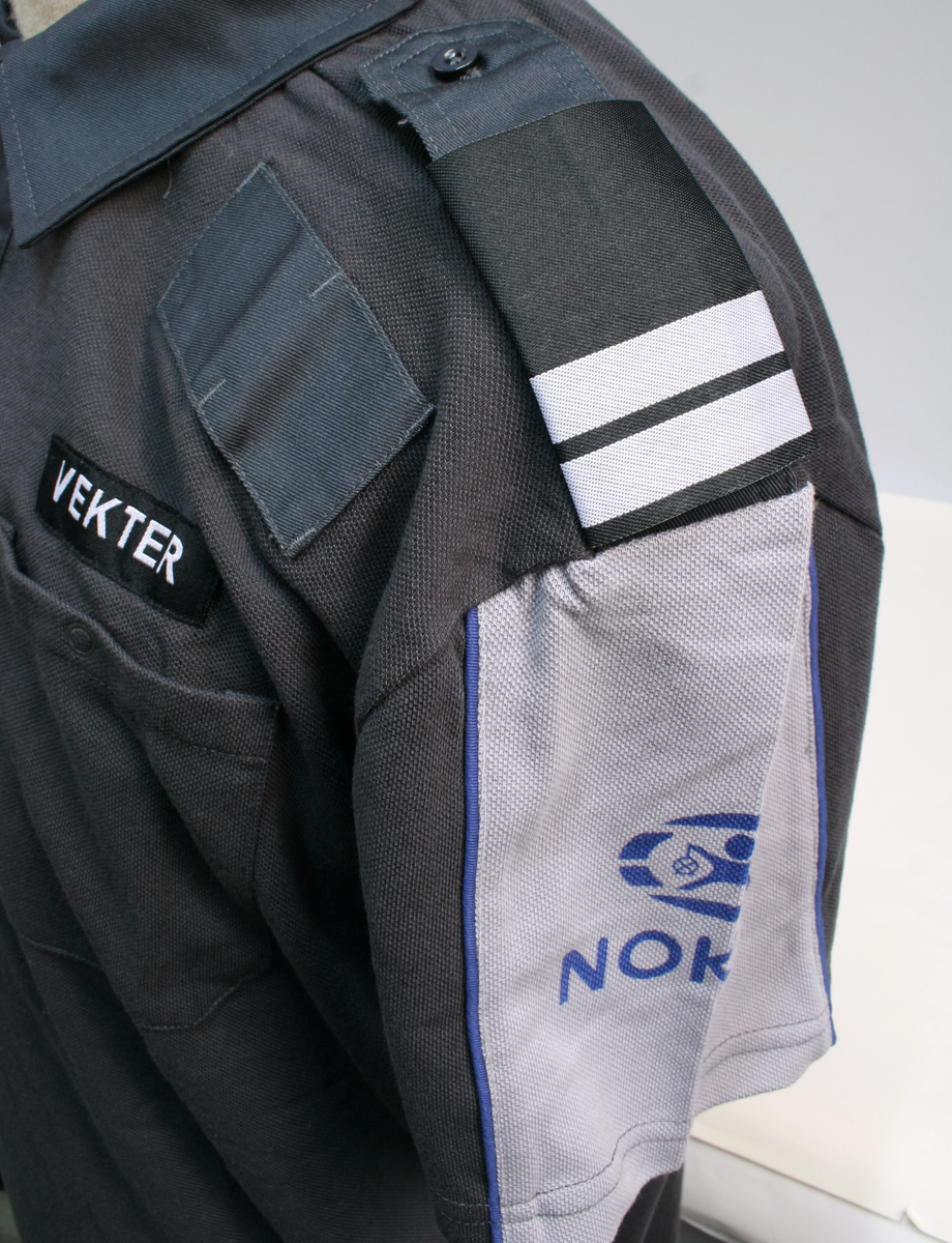 Sort uniform bestående av kortermet poloskjorte, bukser og skyggelue med Nokas-logo, og tilhørende belte rigget med lommelykt og walkie-talkie.