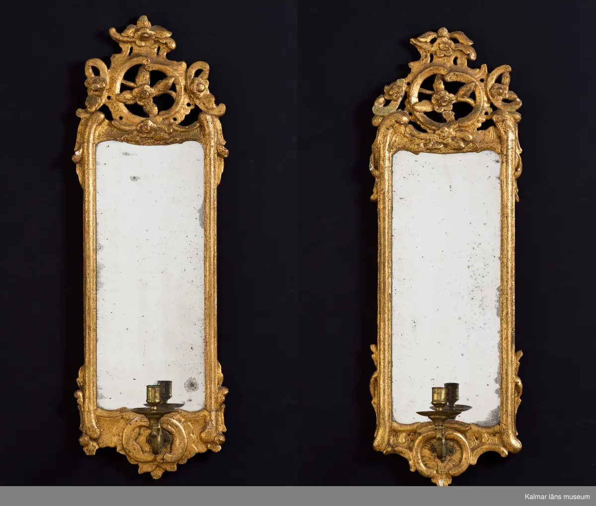 KLM 24949:1-2. Spegellampett, ett par. Guldbronserade ramar av trä med spår efter ursprunglig förgyllning. Rocaille-ornament med tjock kredering överst. Några små olikheter i de båda speglarnas dekor. Otydlig signatur på baksidan. Löstagbara ljuhållare av mässing.