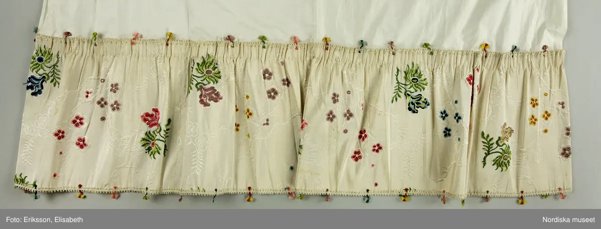 Brudklänning (robe), av broscherat, vitbottnat kinesiskt siden. Nygjord framvåd av fint vitt bomullstyg med kjolvolang nedtill av robens tyg. Två lösa garneringsbårder.