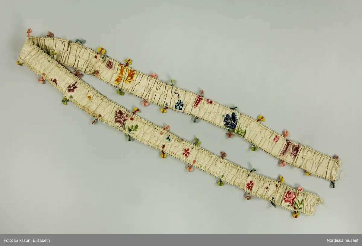 Brudklänning (robe), av broscherat, vitbottnat kinesiskt siden. Nygjord framvåd av fint vitt bomullstyg med kjolvolang nedtill av robens tyg. Två lösa garneringsbårder.