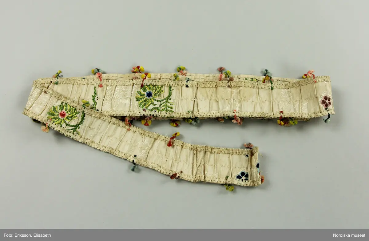 Brudklänning (robe), av broscherat, vitbottnat kinesiskt siden. Nygjord framvåd av fint vitt bomullstyg med kjolvolang nedtill av robens tyg. Två lösa garneringsbårder.