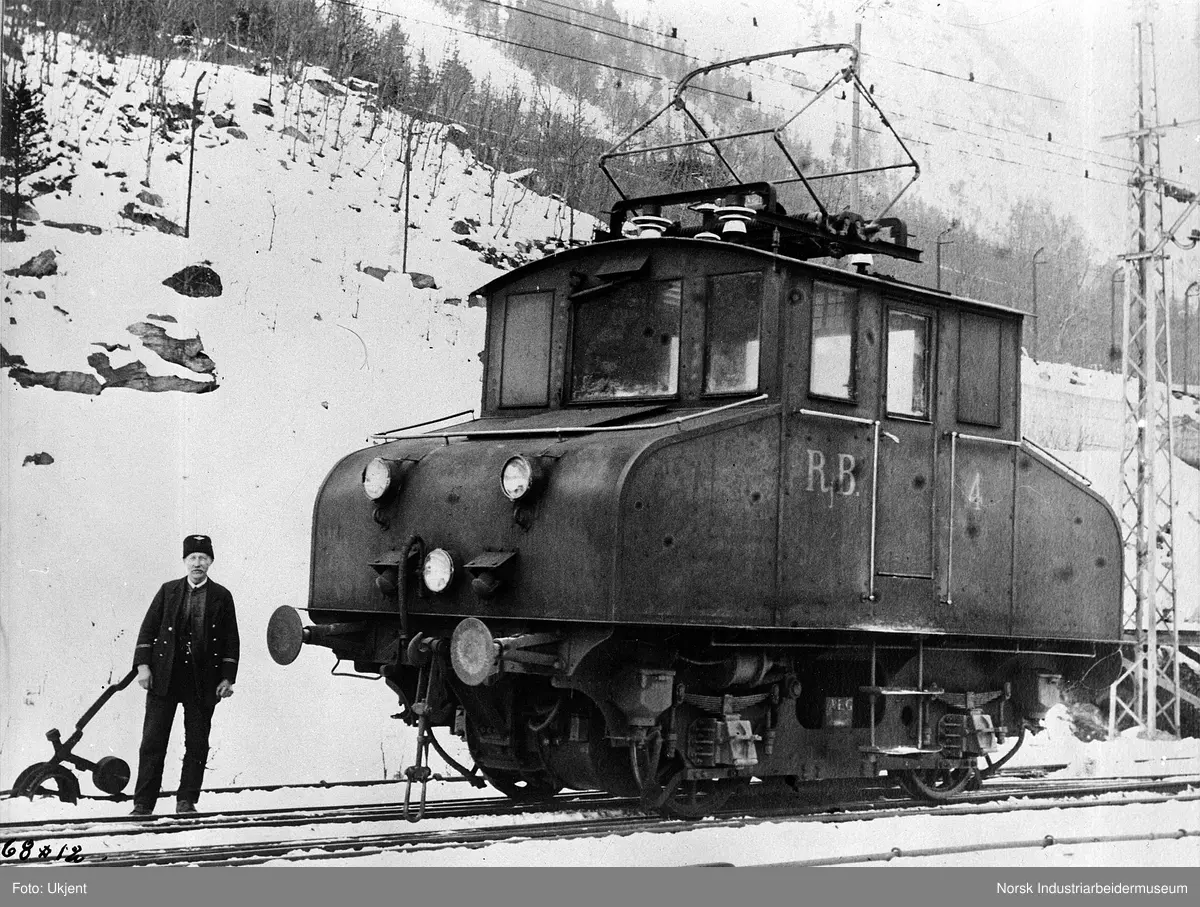Mann kledd i jernbaneuniform stående foran elektrisk lokomotiv RjB 4. Loddstol ved jernbanesporet.