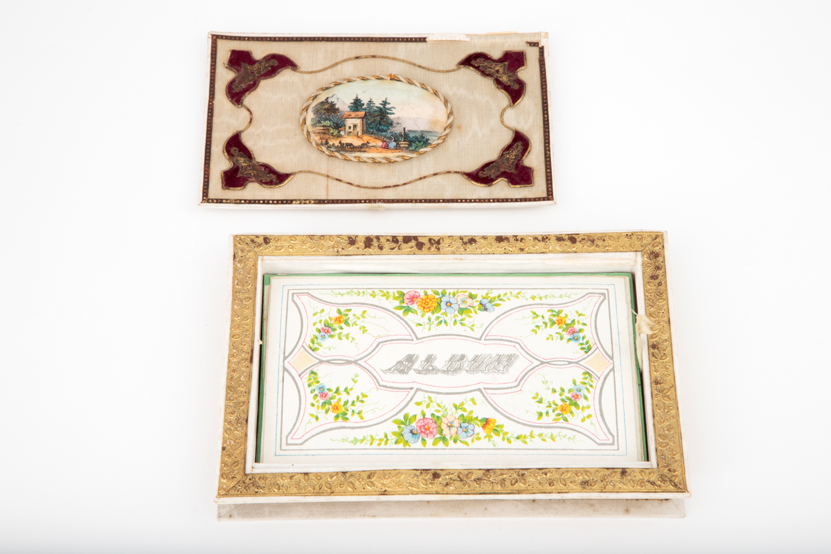 Poesialbum av hvit papp med pålimte gullborder med presset rankemønster. Eskeform med løst løkk som på oversiden er trukket med gulnet hvit rips (stoff), med en medaljong med fargetrykk limt oppå; et landskapsmotiv med kvinner ved en brønn, gjeter og hus. Røde fløyelsapplikasjoner i hjørnene. På lokkets underside er det festet et speil. Under lokket en løs pappplate, hvorpå trykt: ALBUM, omgitt av flerfarget blomsterdekor. Innhold: diverse løse kort (ark) i forskjellige farger, delvis med påskrevne dikt, skrevet cirka 1860.