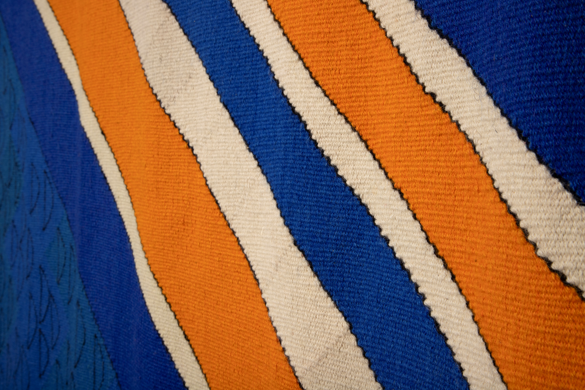 Håndvevd rektangulært billedvev i ull med geometrisk motiv i svart, blått, oransje og beige. Langs venstre og høyre kant er renningstrådene flettet og festet på baksiden med påsydde vevde bånd.