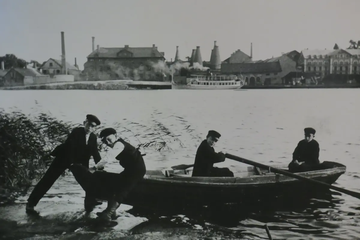 Monterat foto ca 1,5 x 0,8 m visande fyra skolpojkar med vegamössor i en eka med Gustavsbergs fabriker och en ångbåt i bagrunden