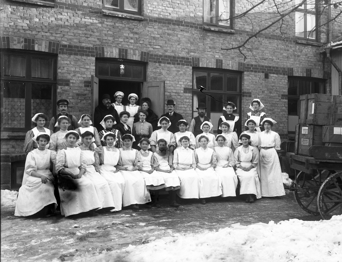 Personalen på Knutsons Marmeladfabrik uppställd ute på innergården i snön. Till höger en vagn med packade produkter. Med på bilden finns: sannolikt fabrikörsparet Aron och Sofie Knutson, frk. Håkansson, kontorist Signhild Pettersson, frk. Knutsson, handelsresande Hertzberg, maskinist Pettersson, gårdskarl Lars Larsson, Emma Johansson, Edit Andrén, Elin Berglund, Josefina Johansson, Dina Holmström, Ida Sjölin, Anna Andersson, Augusta Andersson, Augusta Gustafsson, Naemi Nilsson, Anna Andersson, Elin Karlsson, Märta Svensson, Ida Hallberg, Augusta Möller, Märta Möller, Julia Johansson, Anna Johansson, Elsa Andersson, Sara Nordin. Fabriken låg då i kv Flickskolan.
