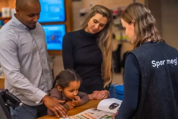 To vaksne og eitt barn ser i eit hefte saman med ein person som har teksten Spør meg! på ryggen. 