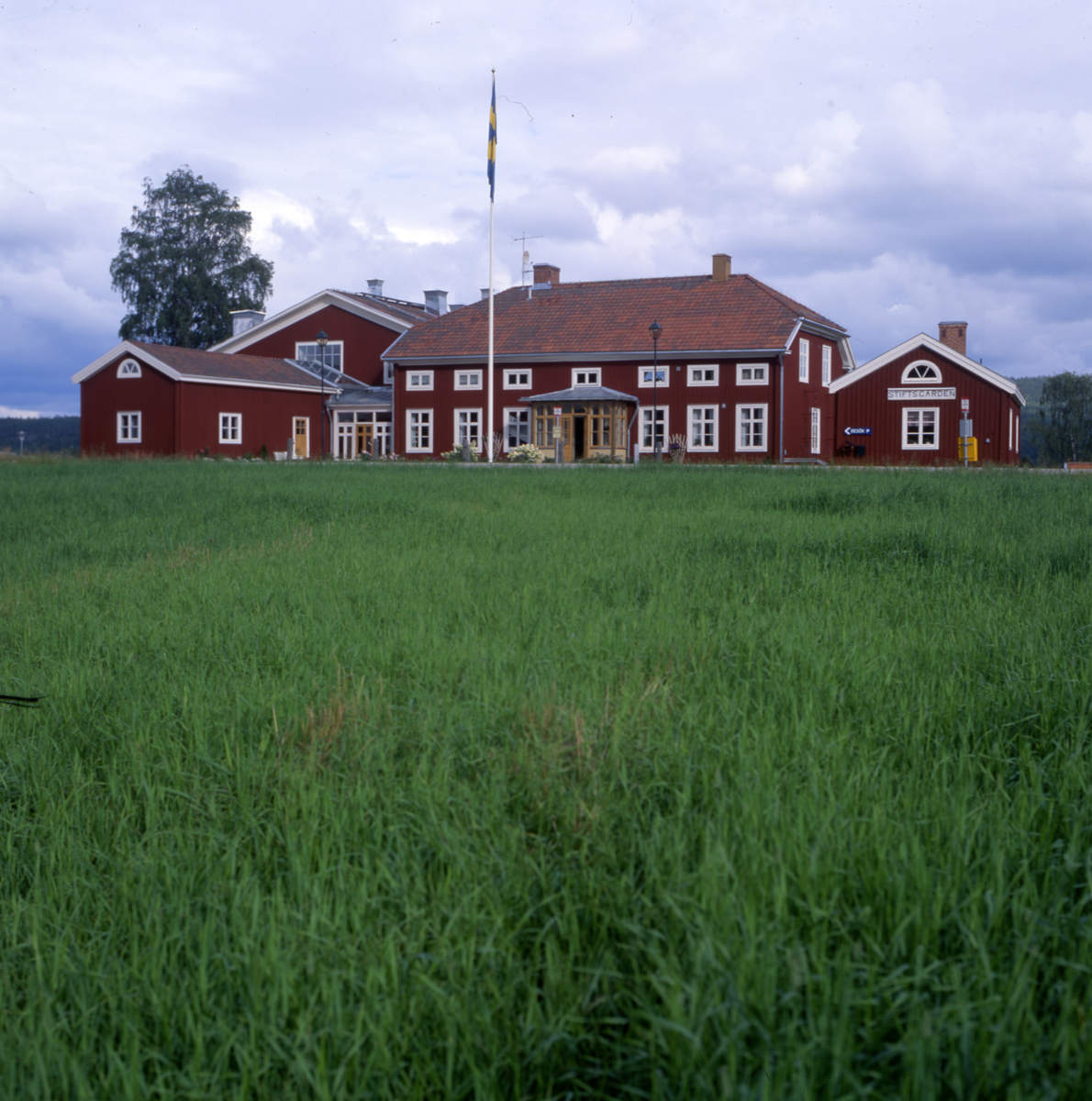 Gård med flera byggnader, glasveranda och flaggstång. Framför gården är marken gräsbevuxen.