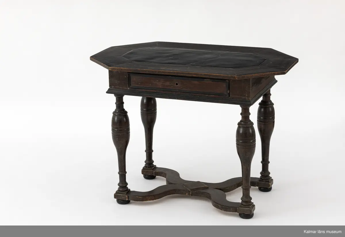 KLM 9107. Bord. Av furu med åttakantig träskiva som i mitten är försedd med en inlagd skifferskiva. På långsidan en låda. Lådans knopp eller handtag saknas. Fyra svarvade ben som nedtill är förbundna med en krysslå. Målad i tunn brun färg, kulfötter och lister i svart.