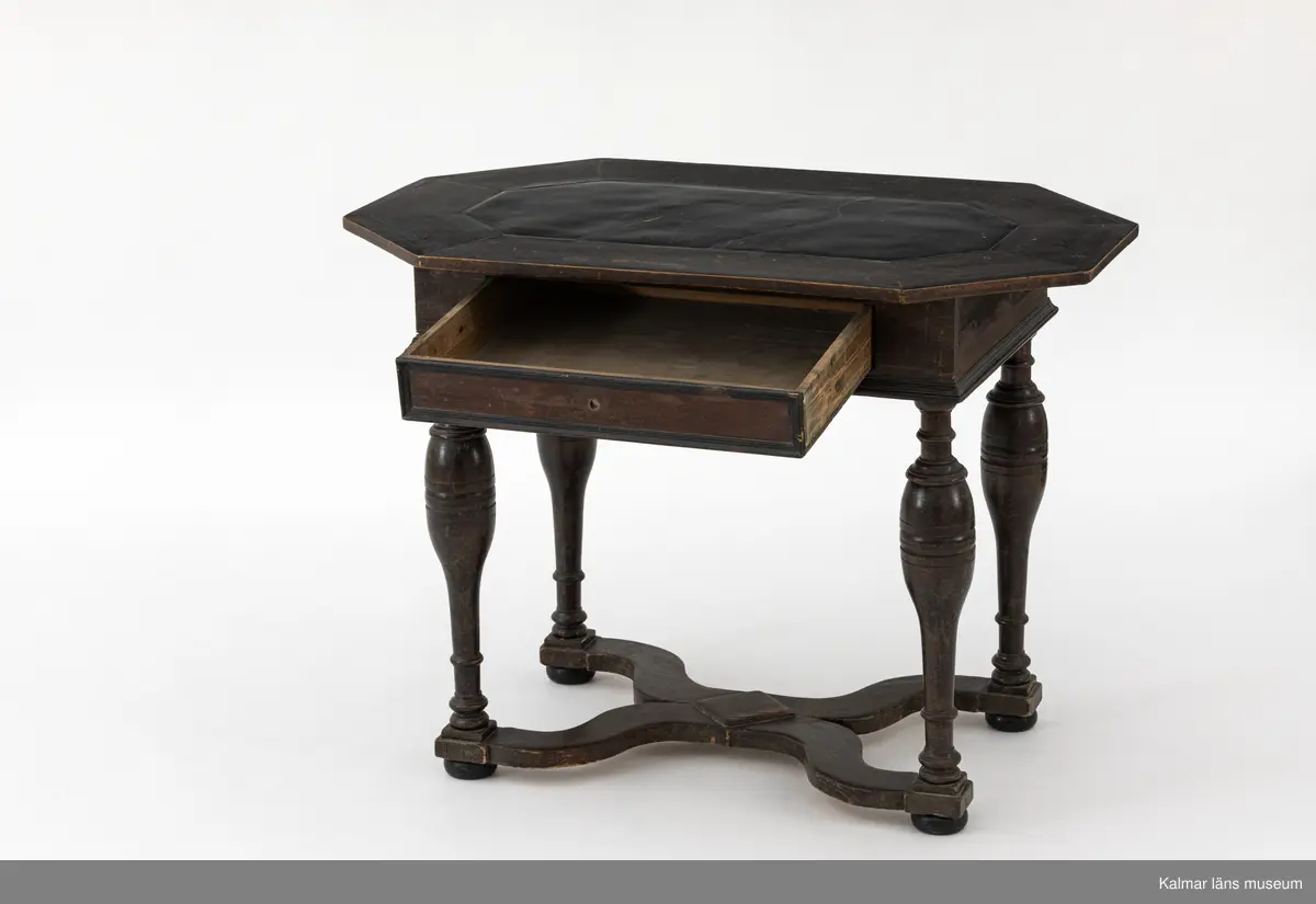 KLM 9107. Bord. Av furu med åttakantig träskiva som i mitten är försedd med en inlagd skifferskiva. På långsidan en låda. Lådans knopp eller handtag saknas. Fyra svarvade ben som nedtill är förbundna med en krysslå. Målad i tunn brun färg, kulfötter och lister i svart.