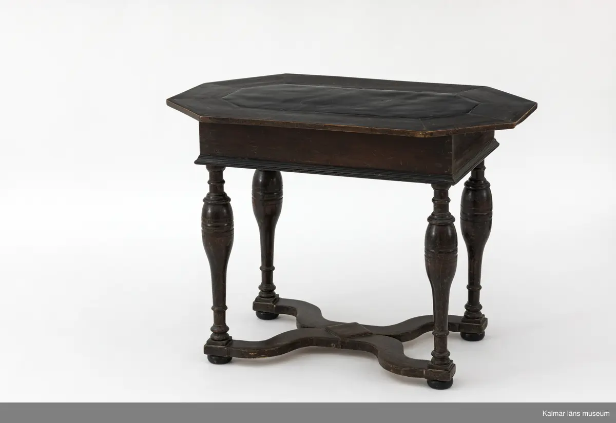 KLM 9107. Bord. Av furu med åttakantig träskiva som i mitten är försedd med en inlagd skifferskiva. På långsidan en låda. Lådans knopp eller handtag saknas. Fyra svarvade ben som nedtill är förbundna med en krysslå. Målad i tunn brun färg, kulfötter och lister i svart.