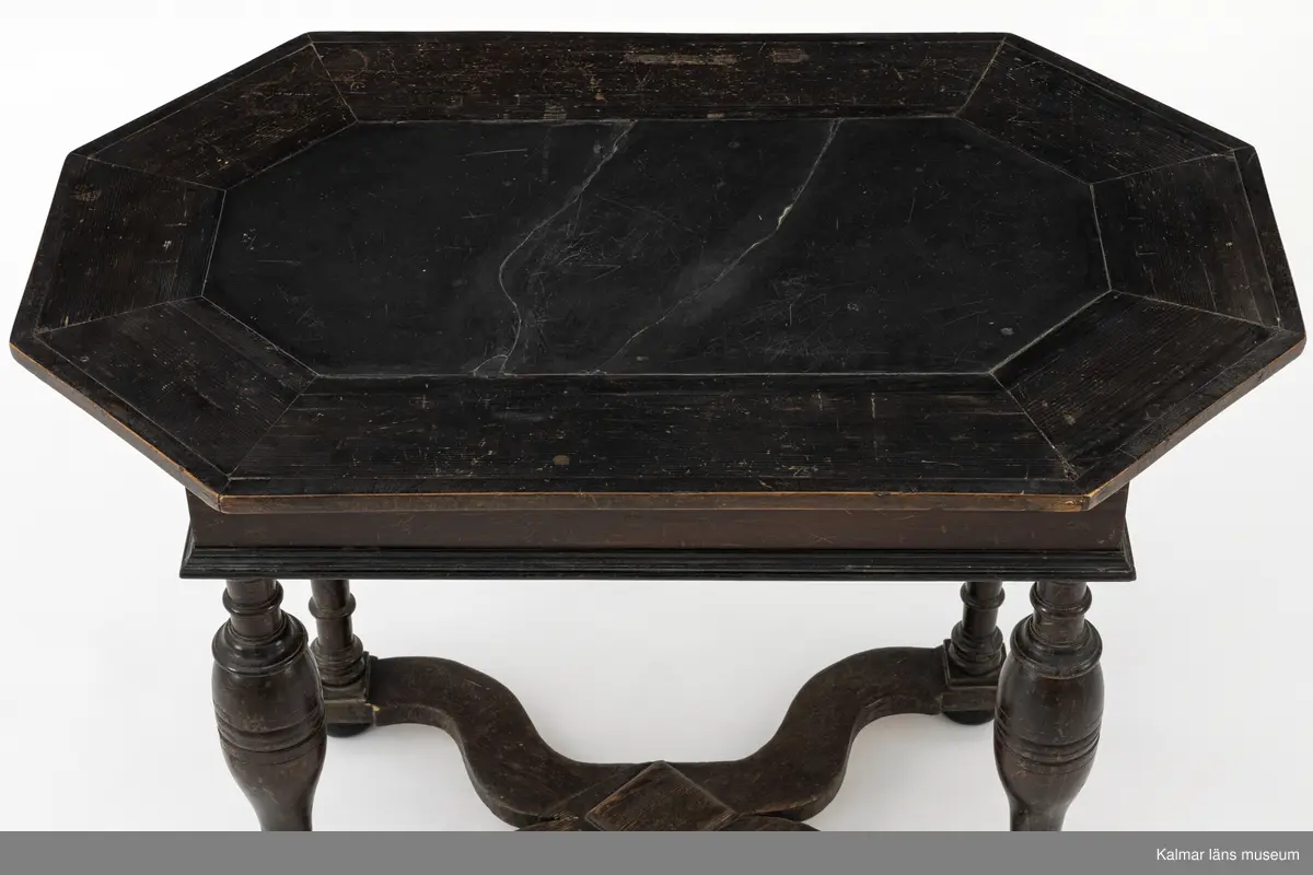 KLM 9107. Bord. Av furu med åttakantig träskiva som i mitten är försedd med en inlagd skifferskiva. På långsidan en låda. Lådans knopp eller handtag saknas. Fyra svarvade ben som nedtill är förbundna med en krysslå. Målad i tunn brun färg, kulfötter och lister i svart.