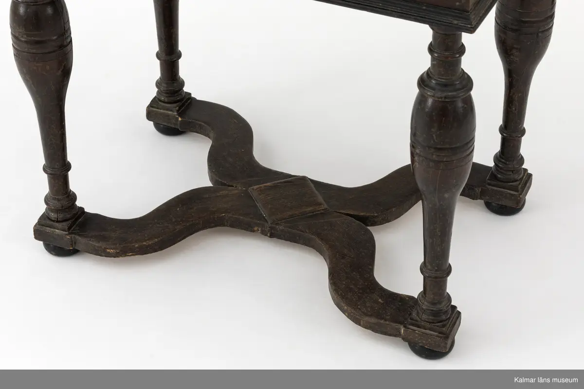 KLM 9107. Bord. Av furu med åttakantig träskiva som i mitten är försedd med en inlagd skifferskiva. På långsidan en låda. Lådans knopp eller handtag saknas. Fyra svarvade ben som nedtill är förbundna med en krysslå. Målad i tunn brun färg, kulfötter och lister i svart.