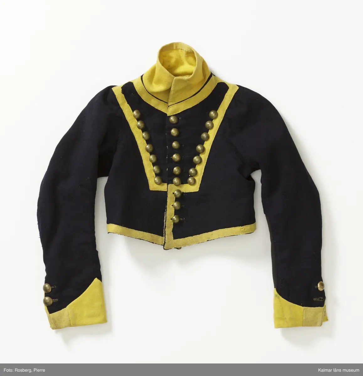 KLM 14853:1. Jacka. Kollett. Till fanjunkareuniform, barnstorlek. Smålands dragoner. Kort jacka av mörkblått kypertvävt ylle med upprättstående ofodrad krage. Krage  ärmuppslag och kanter i gult kläde. Knäpps i en rad med nio st. blanka kulknappar av gulmetall, fästa med grov tråd innanför fodret. På livet fram fastsytt gult band i V-form. Längs bandet sju st. prydnadsknappar av gulmetall. Små skört som står ut mitt bak. Ärmuppslagen är sydda, dekorativa, ej vikta. Ärmar knäpps i nederkant med två st. knappar. Vänster ärm har svart tyg på baksidan, ev. en sentida lagning. Innerfoder i gul boj, ärmfoder i linne. Yttertyget skarvat, bl.a. i ärmslut.