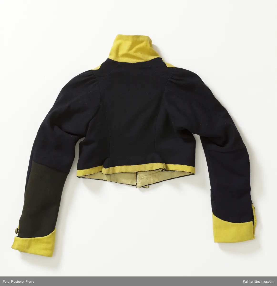 KLM 14853:1. Jacka. Kollett. Till fanjunkareuniform, barnstorlek. Smålands dragoner. Kort jacka av mörkblått kypertvävt ylle med upprättstående ofodrad krage. Krage  ärmuppslag och kanter i gult kläde. Knäpps i en rad med nio st. blanka kulknappar av gulmetall, fästa med grov tråd innanför fodret. På livet fram fastsytt gult band i V-form. Längs bandet sju st. prydnadsknappar av gulmetall. Små skört som står ut mitt bak. Ärmuppslagen är sydda, dekorativa, ej vikta. Ärmar knäpps i nederkant med två st. knappar. Vänster ärm har svart tyg på baksidan, ev. en sentida lagning. Innerfoder i gul boj, ärmfoder i linne. Yttertyget skarvat, bl.a. i ärmslut.