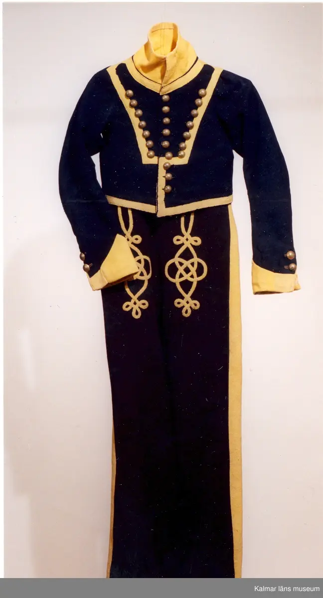 KLM 14853:1. Jacka. Kollett. Till fanjunkareuniform, barnstorlek. Smålands dragoner. Kort jacka av mörkblått kypertvävt ylle med upprättstående ofodrad krage. Krage  ärmuppslag och kanter i gult kläde. Knäpps i en rad med nio st. blanka kulknappar av gulmetall, fästa med grov tråd innanför fodret. På livet fram fastsytt gult band i V-form. Längs bandet sju st. prydnadsknappar av gulmetall. Små skört som står ut mitt bak. Ärmuppslagen är sydda, dekorativa, ej vikta. Ärmar knäpps i nederkant med två st. knappar. Vänster ärm har svart tyg på baksidan, ev. en sentida lagning. Innerfoder i gul boj, ärmfoder i linne. Yttertyget skarvat, bl.a. i ärmslut.