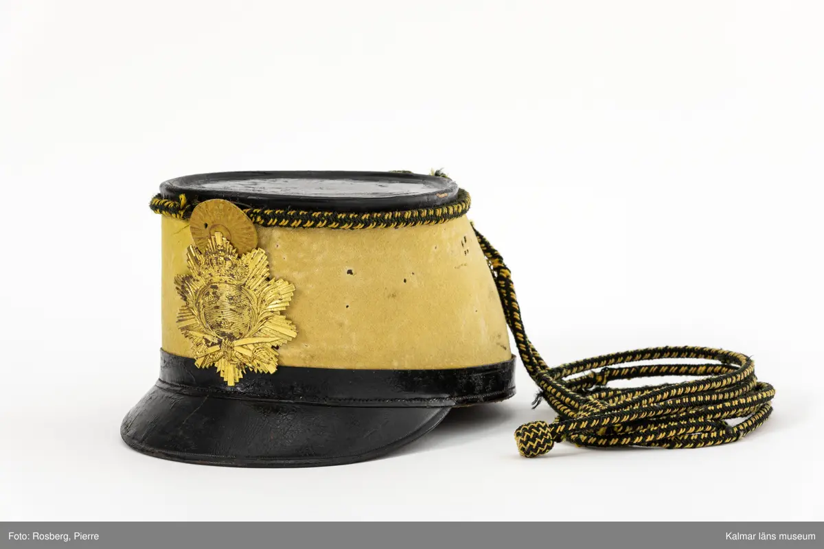 KLM 15169:1. Mössa. Käppi. Husarmössa. Modell 1884. Smålands Husarregemente, Livskvadron. För manskap. Mössa av ljusgult kläde. Rak i sidorna, svagt framåtlutad kulle. Platt ovandel av svartlackat skinn. Skärm fram av svartlackat läder. Längs nederkanten smal remsa av svart skinn, mitt bak fastsatt kil av svart skinn. Vid mössans överkant ett fastsytt flätat snöre av ylle i gult och svart, snöret bildar ögla mitt bak. I öglan fastträdd kordong av lika snöre. Mitt fram gul kokard av läder. Under kokarden mössplåt med lilla riksvapnet, över två st. korsade sablar. Flikklippt svettrem av brunt skinn, öljetterade hål i spetsarna med brunt band trätt igenom. Hakrem, :2, av brunt skinn fastsatt i mössans insidor med spännen av gulmetall, hakremmen knäpps med spänne av gulmetall. På mössans utsida och insida finns totalt fyra röda lacksigill med krona och text; ARME FÖRVALTN.PBOF STÄMPEL.
Hakrem KLM 15169:2 förvaras i mössan.