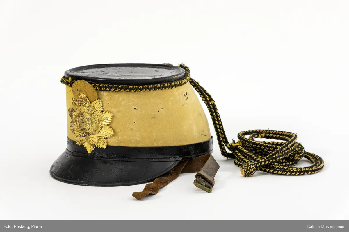 KLM 15169:2. Hakrem. Till Husarmössa. Modell 1884. Smålands Husarregemente, Livskvadron. Rem av brunt läder. Knäpps med spänne av gulmetall och hål, elva st. En hälla av läder vid metallspännet samt en lika hälla som är flyttbar över remmen.
Hakremmen förvaras i mössa KLM 15169:1.