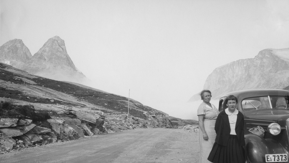 Rast ved veien over Trollstigen mellom Romsdalen og Valldal hvor mor og datter står ved familiens Chevrolet 1936-modell. I bakgrunnen fjellene Kongen, Bispen og Trollklørn.