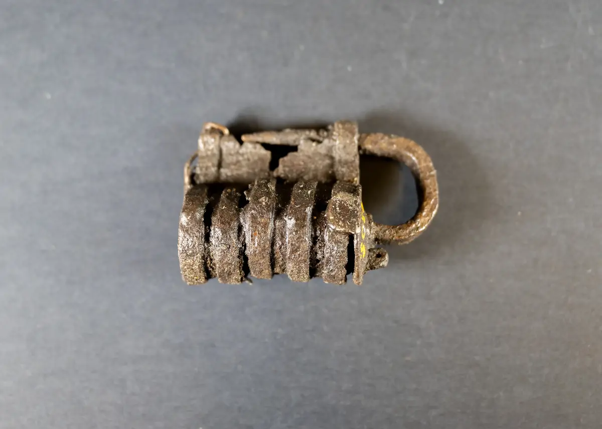 Låshuset cylindriskt med utanpåliggande ringar. Hängbult och regelskiva intakta, nyckelhål i gaveln. Låset har varit i två delar men är lagat i senare tid (vid konserveringen?) med en (koppar)tråd
Av järn, 3 tum långt, avbrutet på mitten
