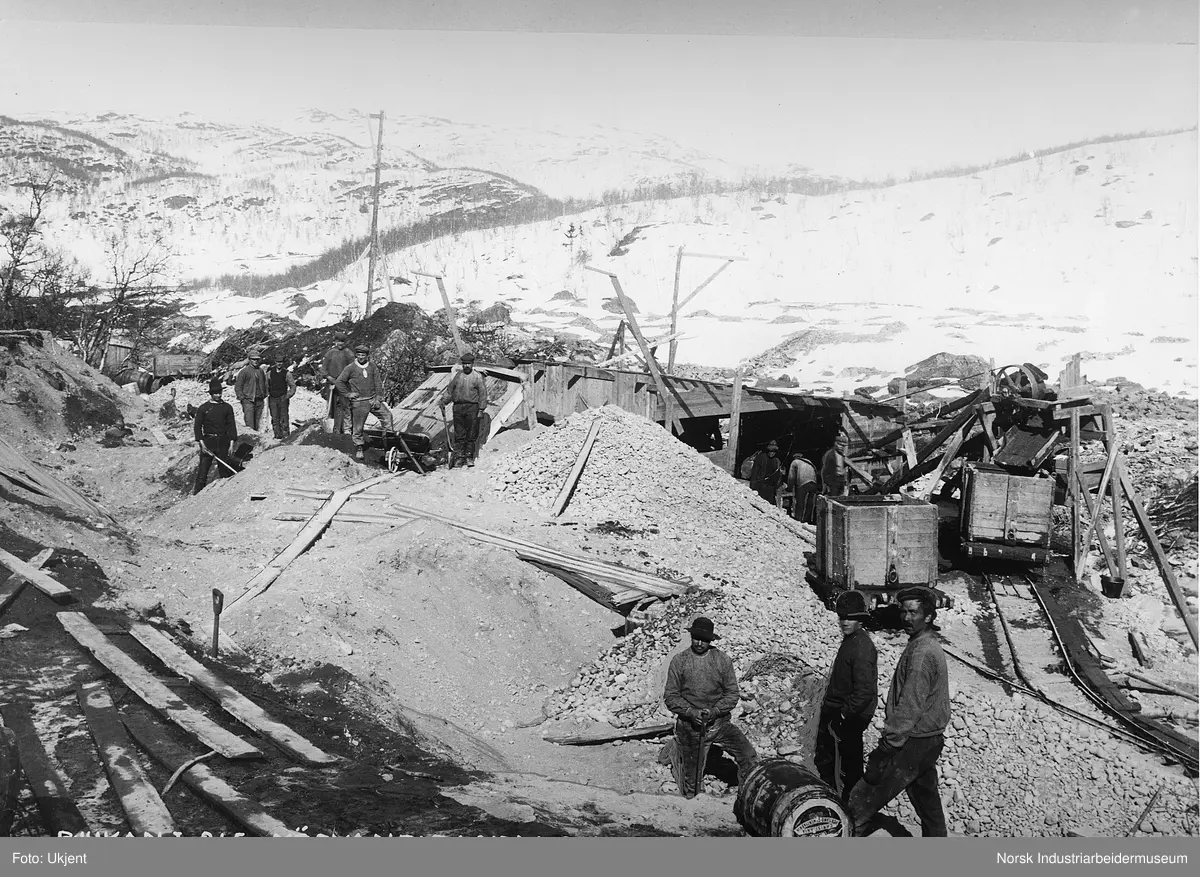 Anleggsarbeidere i sandtak på Møsvann. Smalsporet jernbanespor på området og vogner for sandtransport. Transportbånd for grus fra bygg og opp i vogner.