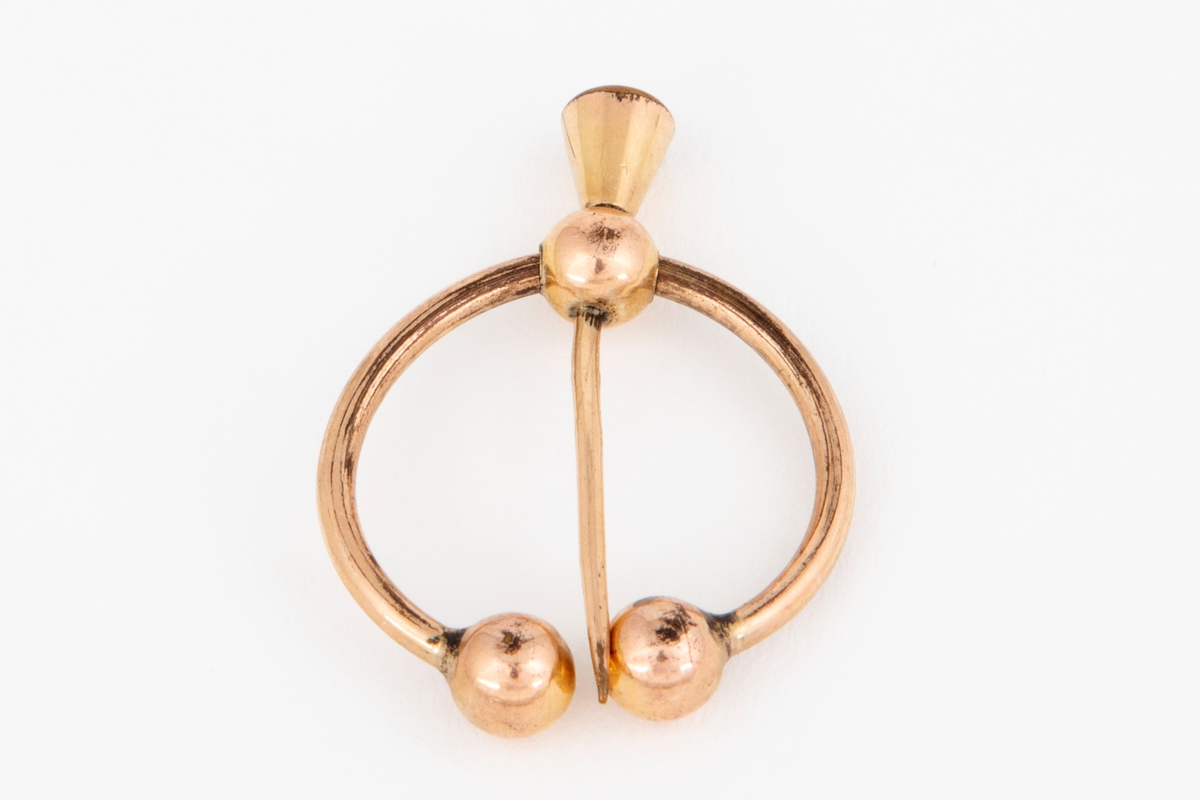 Brosje i gulldublé formet som en åpen ring med to kuler i hver ende. En bevegelig nål med kjegleformet hode er festet til ringen med en tilsvarende kule.