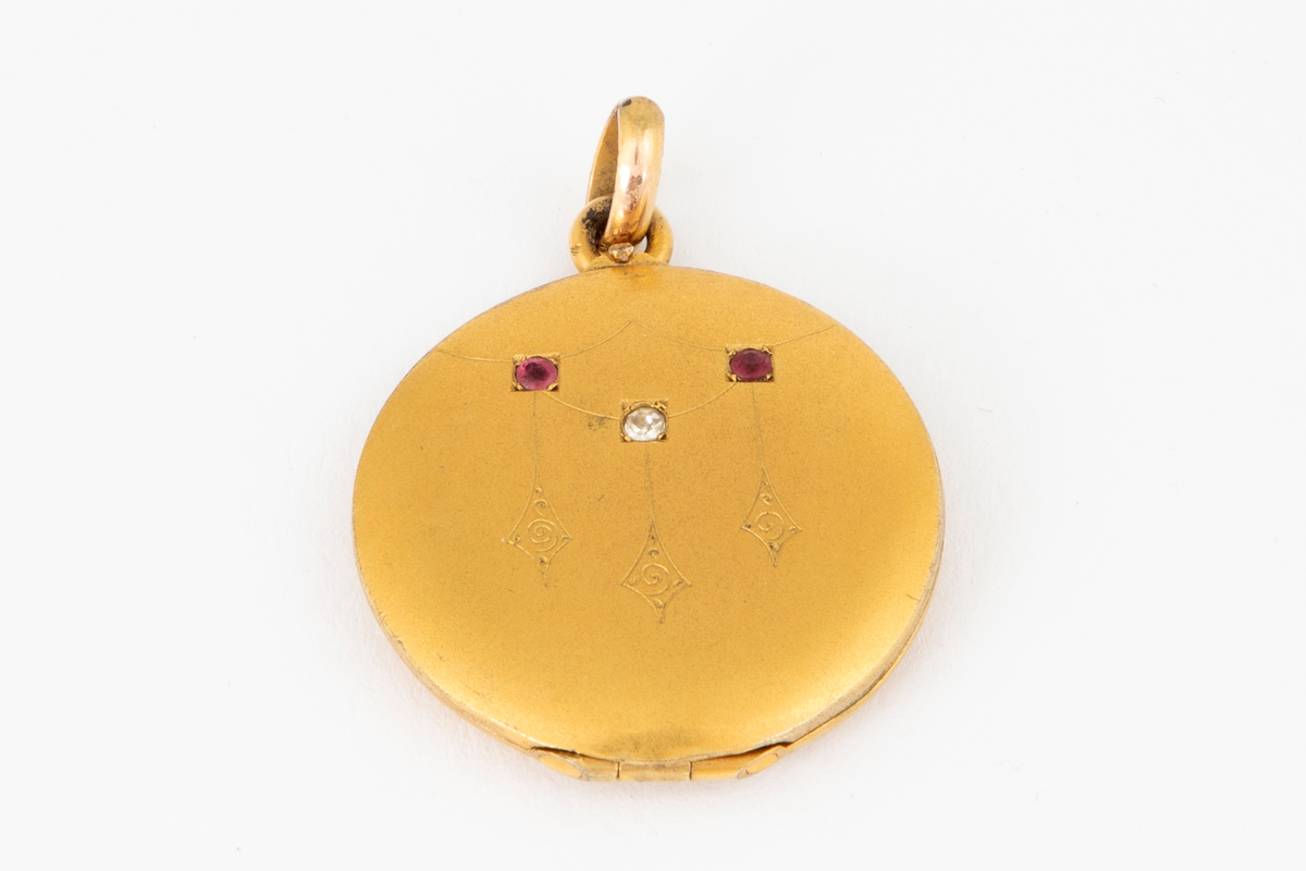 Anheng/ medaljong i forgylt metall med gravert dekor og innlagte stener på forsiden; to røde og en hvit. Glatt bakside. Medaljongen kan åpnes og har rom for to miniatyrbilder eller fotografier bak transparent plast.