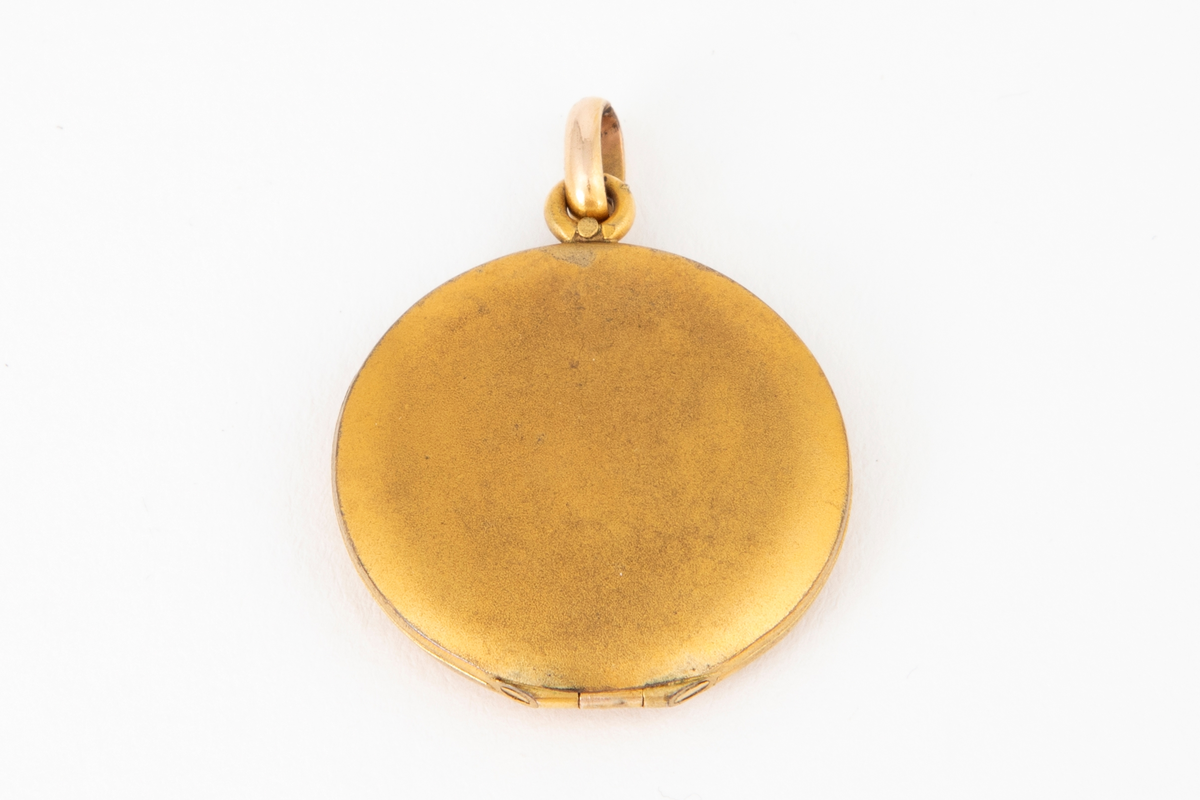 Anheng/ medaljong i forgylt metall med gravert dekor og innlagte stener på forsiden; to røde og en hvit. Glatt bakside. Medaljongen kan åpnes og har rom for to miniatyrbilder eller fotografier bak transparent plast.