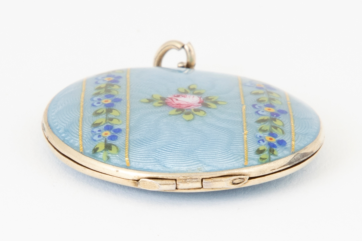 Rundt anheng/ medaljong i forgylt metall med lyseblå emalje over guillochering. Blomsterdekor i rosa, mørkeblå, gul og grønn. Medaljongen kan åpnes og inneholder to svart-hvitt fotografier av en mann.