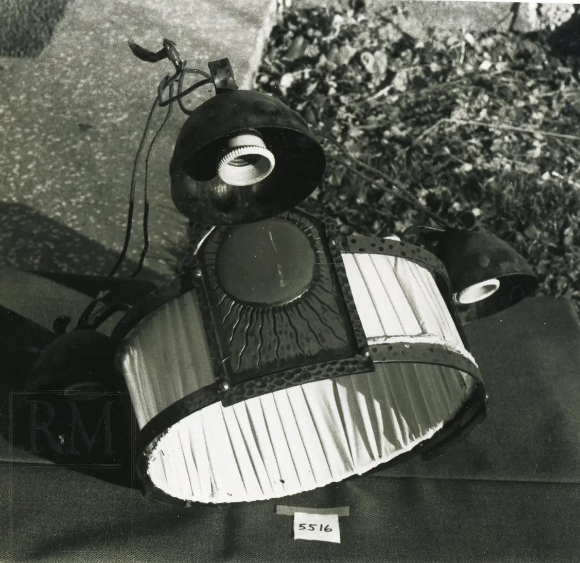 3 lyspærer som henger utover lampens base. Fra de 3 lyspæreholderne kommer ledningstråd som treffer sammen på toppen. Hoveddelen av lampen er rund  med en nylon skjerm. Lyspæreholder av bakkolitt. Hvert lys står på et "skjold" som er en del av (basen) bunnen.