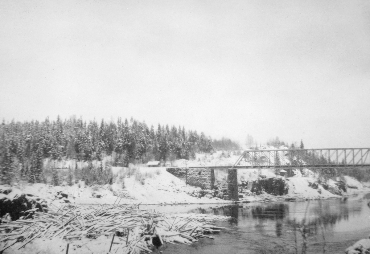 Elvelandskap ved Kjelåsskjæret i Askim i Østfold, fotografert vinteren 1919-20.  Kjelåsskjæret var en liten holme ved innløpet til det øverste fallet i Fossumfossen, et fall som også ble kalt Bjørkeskjærfossen.  Ved denne holmen hadde det lett for å danne seg vaser av fløtingstømmer, noe som vises tydelig på dette bildet, der det ligger en haug av tett sammenpressete, sprikende og til dels snødekte tømmerstokker og danner en propp i det ellers isfrie elveleiet.  Ovenfor, til høyre i bildet, ser vi ei jernbanebru av støpejernselementer som krysser elva i ett spenn.  Brua kviler på landkar av hoggen gråstein.  Like inntil brukaret på vestre side skimter vi taket på en liten bygning.  Bakenfor ser vi en bergrygg med granskog. 