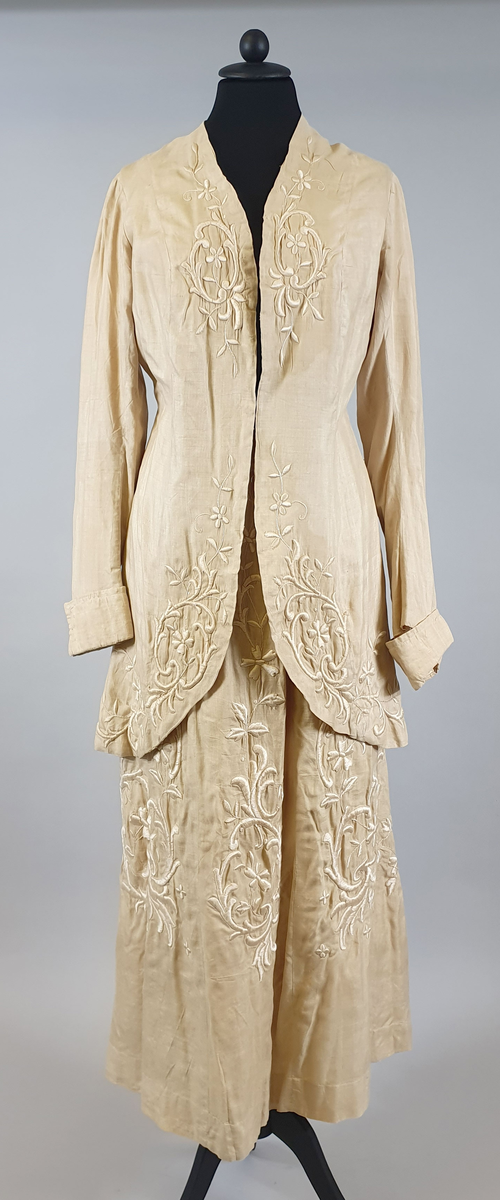 Naturfarget drakt av lin, fôret med silke, bestående av jakke og skjørt. Det følger også med et underskjørt av silke. Jakken er buet over brystet og nederst foran og er innsvinget i livet, og har lange ermer med mansjetter, og har engelsk broderi både på forsiden og på ryggen. Skjørtet har samme type broderi som jakken,