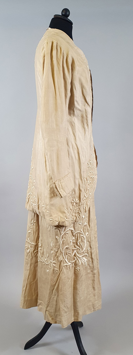 Naturfarget drakt av lin, fôret med silke, bestående av jakke og skjørt. Det følger også med et underskjørt av silke. Jakken er buet over brystet og nederst foran og er innsvinget i livet, og har lange ermer med mansjetter, og har engelsk broderi både på forsiden og på ryggen. Skjørtet har samme type broderi som jakken,